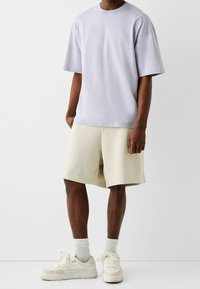 Světle levandulové oversized tričko, béžové kraťasy a krémové tenisky s bílými ponožkami. Měkká bavlněná látka, jednoduchý design, žádné viditelné vzory.