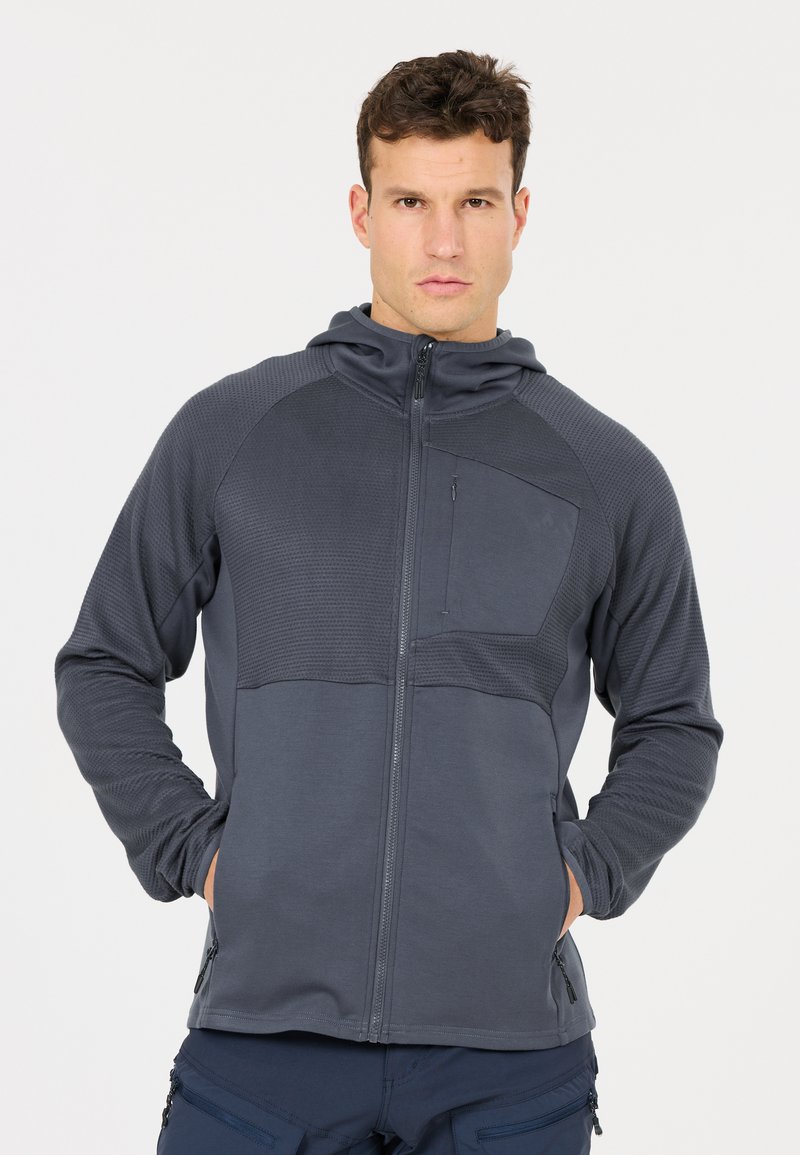 Man draagt een donkergrijze zip-up hoodie met gestructureerde stof op de schouders en borstzak, handen in de zijzakken, tegen een effen achtergrond.