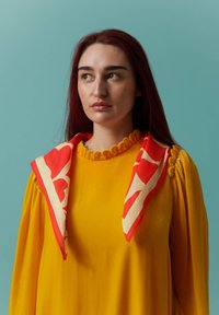 Femme aux longs cheveux roux portant une robe jaune moutarde et une écharpe à motifs rouge et blanc, regardant légèrement sur sa gauche devant un fond bleu-vert.