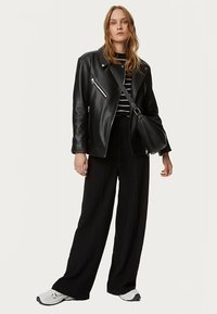 Giacca da motociclista in pelle nera con cerniere argentate, top a righe bianche e nere, pantaloni neri larghi e scarpe da ginnastica bianche, con una borsa a tracolla nera.