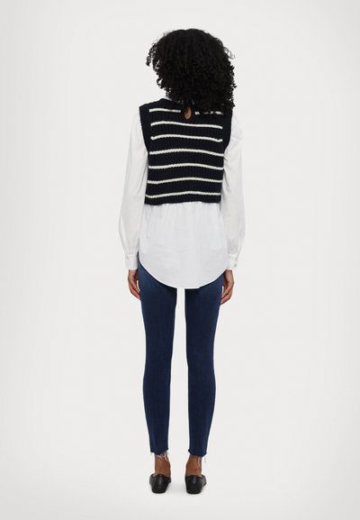 Kötött, rövidített mellény navy és fehér csíkokkal, fehér ing fölött viselve. Slim fit farmernadrág, kifröcskölt széllel és fekete lapos cipővel.