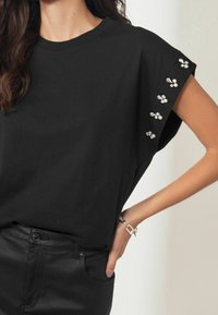 Czarny T-shirt z krótkim rękawem o luźnym kroju, z ozdobami z pereł i kryształów na rękawach. Miękka tkanina, casualowy design.