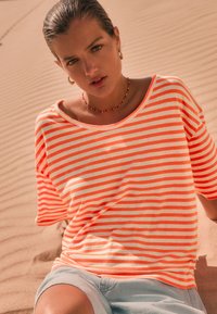 Striped oversized t-shirt in bright orange and white, made of soft fabric, paired with light blue shorts, set against sandy background.