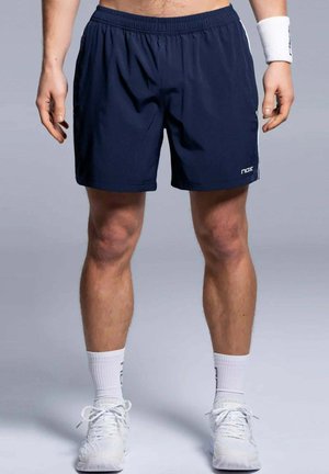 Man draagt marineblauwe sportshorts, witte sportschoenen, witte sokken en een wit polsbandje, staande voor een lichtgrijze achtergrond.