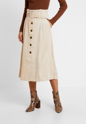 Kvinde iført beige cordnede midi-nederdel med skildpaddemønstrede knapper og matchende bælte, kombineret med brune ankelstøvler med slangeskindsmønster.