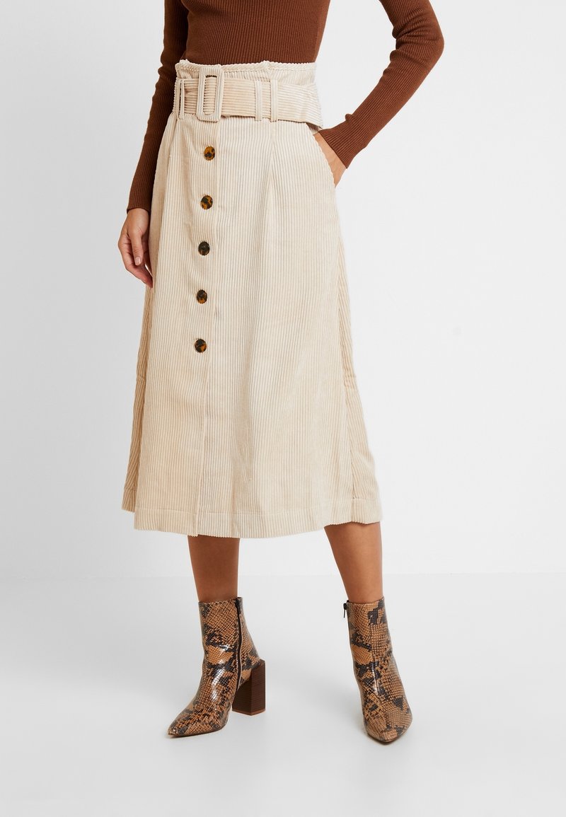 Femme portant une jupe midi beige en velours côtelé avec des boutons en écaille de tortue et une ceinture assortie, associée à des bottines marron à imprimé serpent.