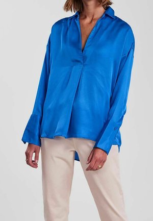 Femme portant un chemisier en satin bleu vif à manches longues et ample, associé à un pantalon beige clair, sur un fond uni.