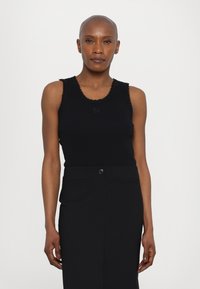 Schwarzes Tanktop mit gewelltem Saum am Ausschnitt und an den Armausschnitten, mit einem gestickten Logo. Kombiniert mit einem schwarz-hoch taillierten Rock.