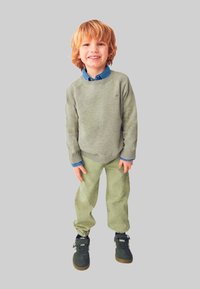 Bambino che indossa un maglione verde chiaro sopra una camicia blu con bottoni, abbinato a pantaloni di velluto a coste verde chiaro e scarpe da ginnastica scure.