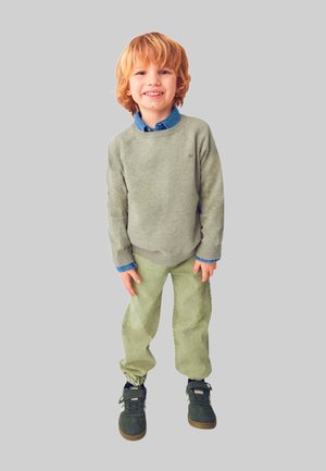 Bambino che indossa un maglione verde chiaro sopra una camicia blu con bottoni, abbinato a pantaloni di velluto a coste verde chiaro e scarpe da ginnastica scure.