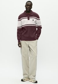 Mand med en bordeaux og hvidstribet langærmet polo sweater, beige brede bukser og lysegrå sneakers, stående med den ene hånd i lommen.