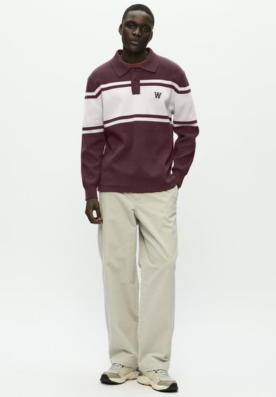 Homme portant un pull polo à manches longues bordeaux et blanches rayé, un pantalon large beige et des baskets gris clair, se tenant avec une main dans la poche.