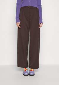 Personne portant un pantalon large marron, un haut à manches longues violet, des chaussures violet clair et des chaussettes florales colorées, debout sur un sol blanc.