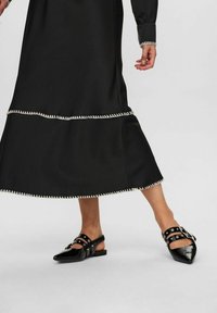 Černé midi šaty s rozšířeným lemem a bílým dekorativním šitím, spárované s lesklými černými balerínami s špičatou špičkou a páskovými akcenty.