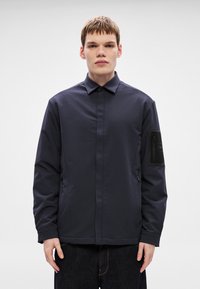 Temno modra jakna s kragnim dizajnom, sprednjim zadrgo in stranskim žepom; na levi rokav ima teksturirano črno šečko.