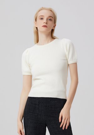 Haut en tricot blanc à manches courtes avec manches bouffantes, col rond et ourlet côtelé. Le tissu semble doux et texturé. Porté avec un pantalon foncé.