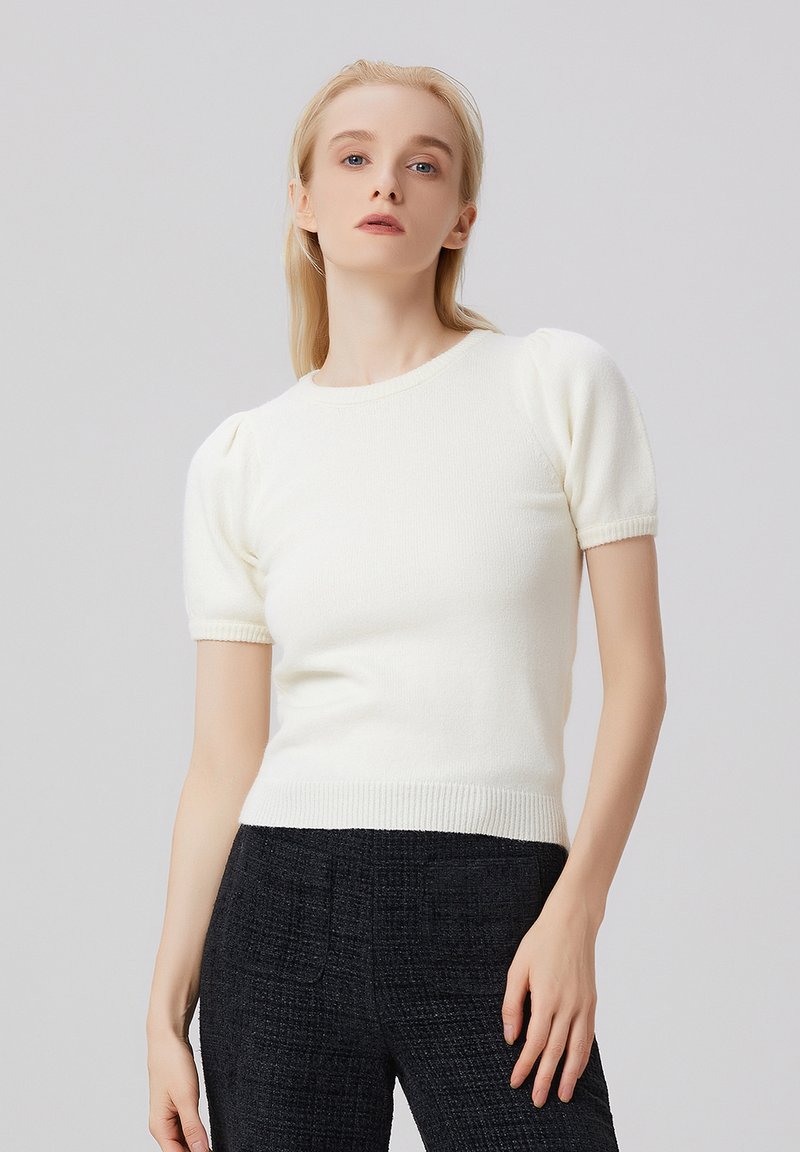 Haut en tricot blanc à manches courtes avec manches bouffantes, col rond et ourlet côtelé. Le tissu semble doux et texturé. Porté avec un pantalon foncé.