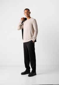 Homme portant un pull beige et un pantalon noir tenant une veste noire sur l'épaule, se tenant devant un fond blanc uni.
