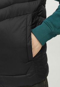 Fekete puffervest steppelt dizájnnal, oldalzsebbel és alulról kikandikáló zöld bordás mandzsettával. Lágy, texturált szövet látható.