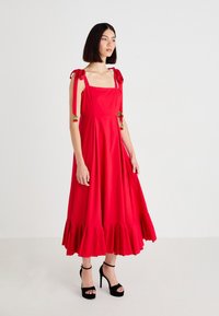 Woman wearing a red midi dress with shoulder ties and cherry embellishments, paired with black high-heeled platform sandals, standing on white floor.