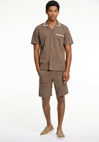 Brun kortærmet skjorte med diskret tekstur og hvid kant, kombineret med matchende shorts. Model iført slip-on sko.