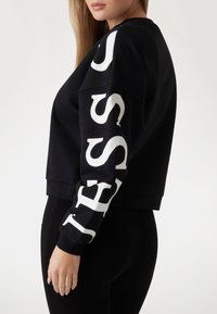 Schwarzer, kürzer geschnittener Sweatshirt mit weißer Aufschrift am linken Ärmel. Hergestellt aus weichem Material, mit einer lockeren Passform und gerippten Bündchen.