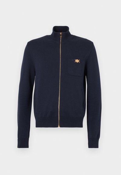 Tamsiai mėlynas megztinis su viso ilgio užtrauktuku, aukštu apykakliu, ribomis rankogaliuose ir maža krūtinės kišene su auksiniu dekoratyviniu sagtimi.