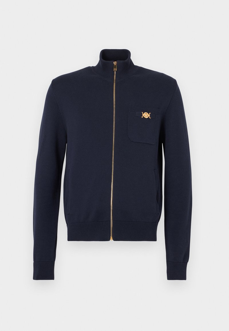 Pull entièrement zippé bleu marine avec col montant, poignets côtelés, et petite poche poitrine ornée d'une fermeture décorative dorée.