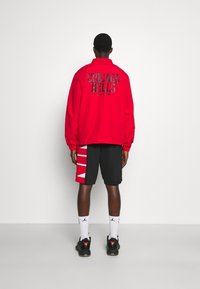 Rode jas met het logo van de "Chicago Bulls" op de rug, gecombineerd met zwarte shorts met rode accenten. Zwarte sportschoenen en witte sokken zichtbaar.