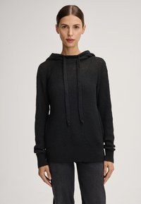 Zwarte gebreide hoodie met een textuurafwerking, lange mouwen, koord in de hals en een relaxte pasvorm. Gecombineerd met donkere broek.