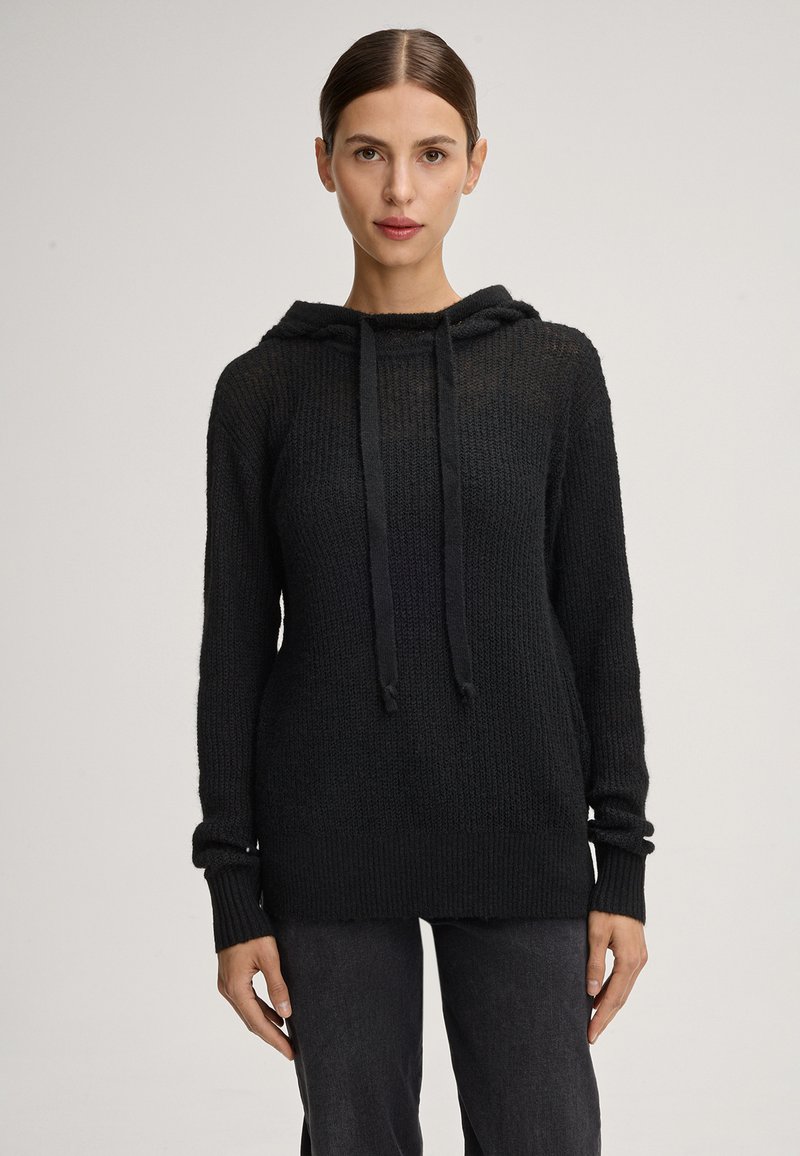 Schwarzer gestrickter Hoodie mit strukturiertem Finish, langen Ärmeln, Kordelzug am Hals und einer lockeren Passform. Kombiniert mit dunklen Hosen.