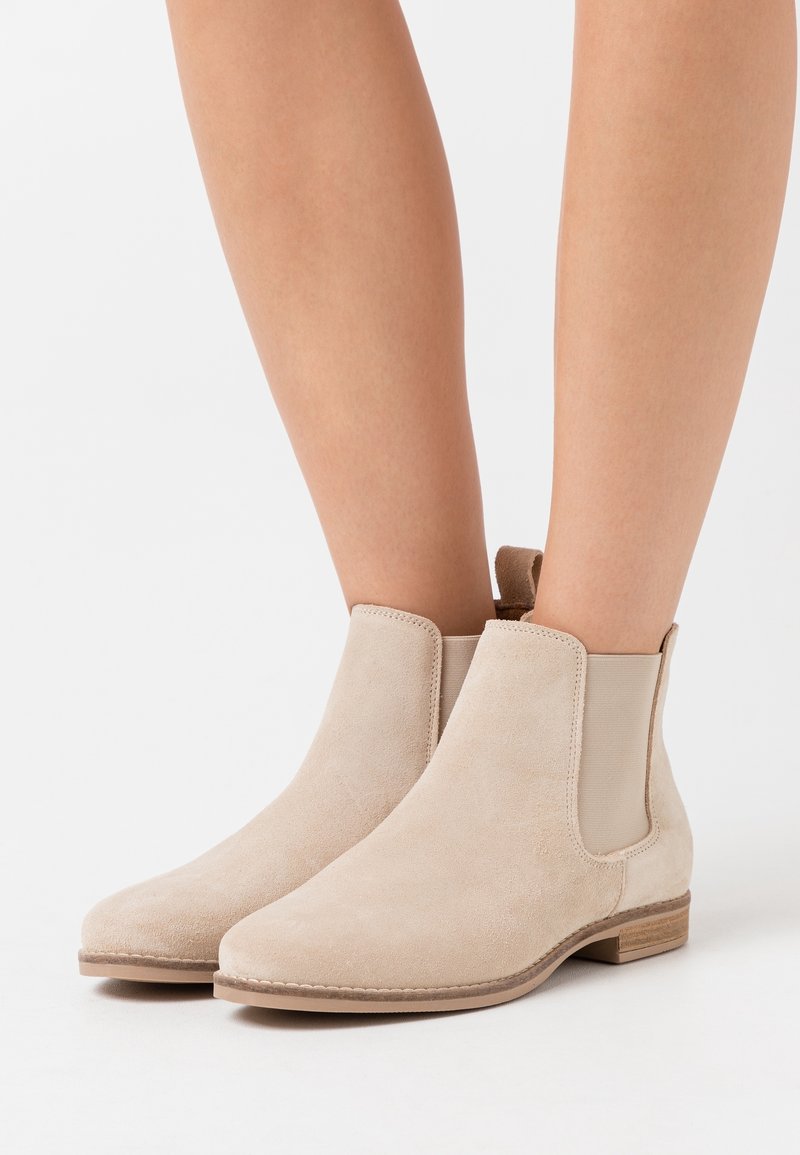 Bottines beiges en daim avec des panneaux latéraux élastiques et des petits talons en bois, portées sur des jambes nues sur fond blanc.