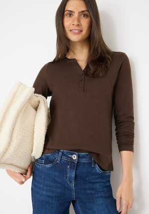 Brun langærmet henley skjorte med knapstolpe, båret med blå jeans, holder en hvid fleecejakke. Glat tekstur og tætsiddende design.