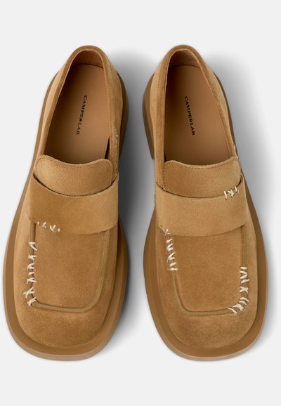 Lysebrune ruskindssko uden snørebånd med hvid dekorativ syning og gummisåler, set oppefra mod en hvid baggrund.