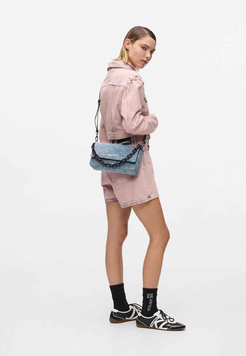 Jeune femme en veste rose et short, portant des chaussettes noires et des baskets, avec un sac à bandoulière bleu à chaîne.
