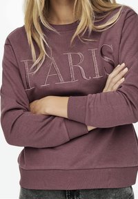 Fialová mikina s vyšitým nápisem "PARIS" v světlejší barvě. Má žebrované manžety a spodní lem, volný střih a měkkou texturu látky.