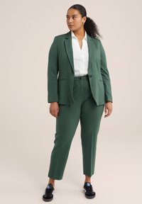 Zelený ušity blazer a odpovídající slim-fit kalhoty vyrobené z hladkého materiálu. V kombinaci s bílou košilí s knoflíky a černými teniskami.