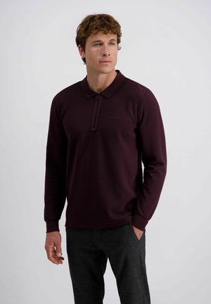 Man staat met zijn linkerhand in zijn zak, gekleed in een donkerpaarse lange mouwen polo en grijs geblokte broek tegen een effen achtergrond.