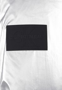 Svart Calvin Klein Jeans-patch med prägling av CK-logotyp på glänsande silverfärgad tygbakgrund.