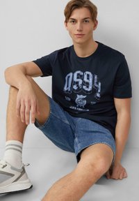 Marineblauwe katoenen t-shirt met grafische print in lichtblauw. Gecombineerd met lichtblauwe denim shorts. Sneakers met gaasaccenten en grijze strepen.