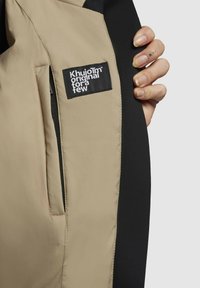 Beige- und schwarze Jacke mit glatter Textur, die ein sichtbares Logo-Patch und eine Reißverschlusstasche auf dem äußeren Stoff aufweist.