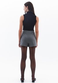 Top sans manches noir associé à un short gris à rayures graphiques taille haute. Le mannequin porte un collant noir transparent et des chaussures à talons bas.