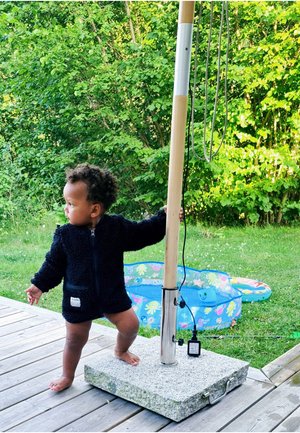 Svart fluffig jacka med dragkedja, stående på en grå stenbas, hållande en träpost med kablar, omgiven av grönska och en liten damm.