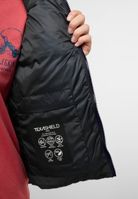 Interno di una giacca nera isolata con una tasca che mostra l'etichetta TEXASHIELD Pro, con icone di windproof, traspirante e repellente all'acqua.