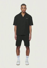 Camiseta preta de manga curta com botões à frente e logotipo, combinada com calções à medida. Usada com meias brancas e ténis. Design simples, tecido leve.