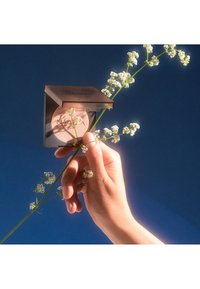 Bronzer compact avec un couvercle en bronze réfléchissant, contenant une poudre rose clair à l'intérieur, posé sur un fond bleu parsemé de délicates fleurs blanches.