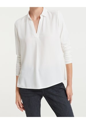 Biała bluzka z długim rękawem, kołnierzykiem i dekoltem w serek, noszona z ciemnoniebieskimi dżinsami na osobie stojącej na jednolitym tle.