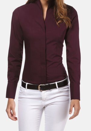 Frau trägt ein eng anliegendes, langärmliges, dunkelviolettes Shirt, das in weiße Hosen mit schwarzem Gürtel gesteckt ist, vor einem einfarbigen hellen Hintergrund.