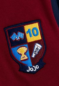 Bordó szövet, hímzett pajzsos folttal, amelyen sakktáblás zászlók, 10-es szám, arany trófea, fehér megafon és 'JoJo' felirat látható.