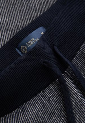 Detail námořnicky modrého žebrovaného pasu se stahovacími šňůrkami na šedobílém pruhovaném materiálu, s modrou značkou „Ombre Heritage".